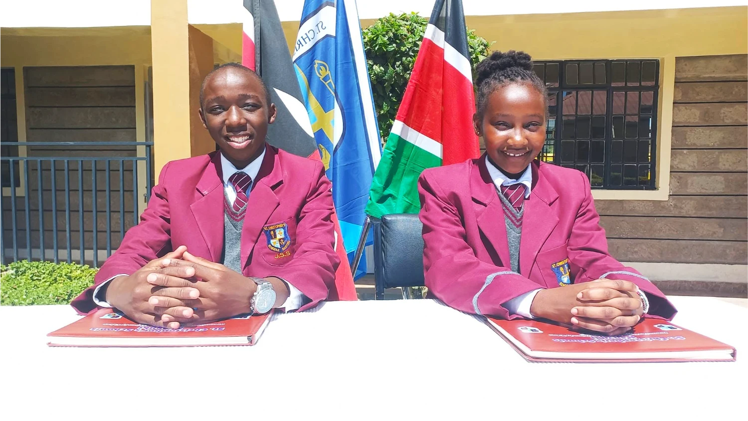 Two St. Christopher's debaters in school blazers seated at the speakers' table in front of the school and Kenyan flags